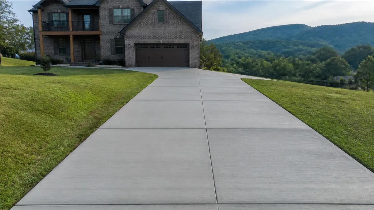 A long new broom-finished concrete driveway with straight saw-cut control joints curving up a hillside lot to a brick two-story home, with wooded ridges behind