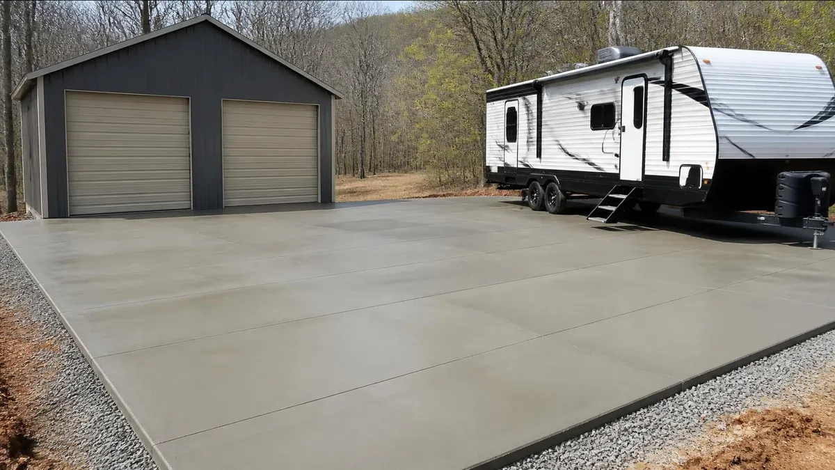A large new concrete pad with saw-cut joints and a gravel edge in front of a two-bay detached garage, with a travel trailer parked on it