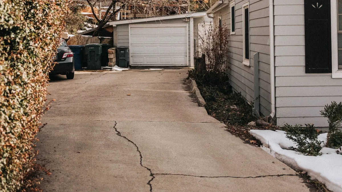 An older concrete driveway beside a house with long cracks running across the slab, old snow along its edge and a detached garage at the far end