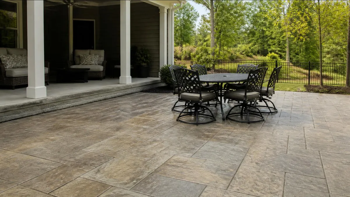 A stamped concrete patio in a gray and tan ashlar slate pattern beside a covered back porch, with a dining set and a wooded yard beyond